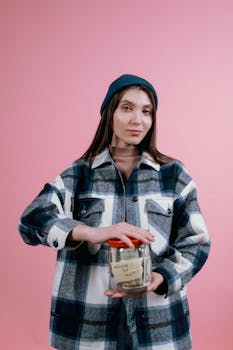 A woman in a plaid shirt and beanie holds a glass jar labeled savings against a pink background.