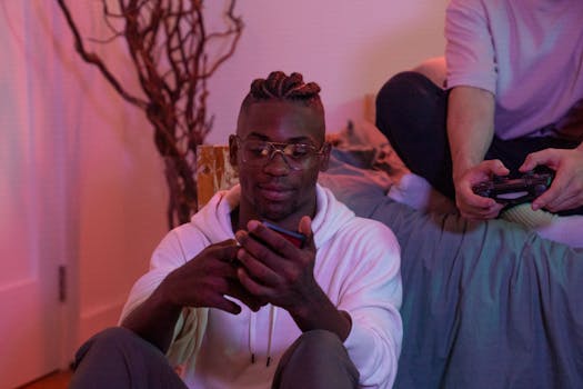 Two friends enjoying a gaming session at home, one using a mobile phone and the other a game controller.