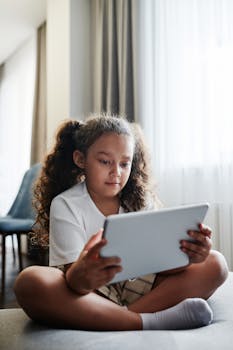Young girl with curly hair using a tablet indoors, sitting cross-legged.