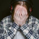 A young gamer covers face with hands in frustration, wearing headphones and plaid shirt in a dimly lit room.
