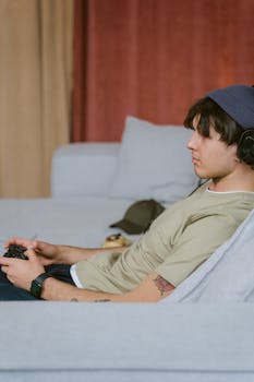 Adult male relaxing on sofa, focused on video gaming with headset and controller.