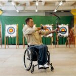 Focused young man in a wheelchair practicing archery indoors, aiming with determination at a target.