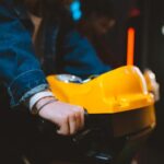 Hand holding handlebar of a yellow arcade racing game machine in a dimly lit setting.
