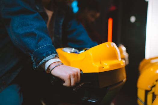Hand holding handlebar of a yellow arcade racing game machine in a dimly lit setting.