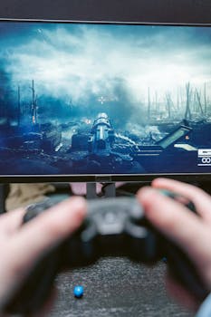 Hands gripping a game controller with action displayed on a monitor in the background.